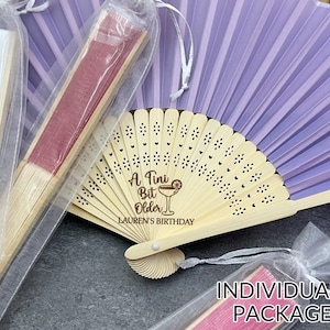 May include: A collection of personalized fans, some in clear packaging. One fan is open, displaying a light wood frame and lavender paper. The open fan has the text "A Tiny Bit Older LAUREN'S BIRTHDAY" and a cocktail graphic.