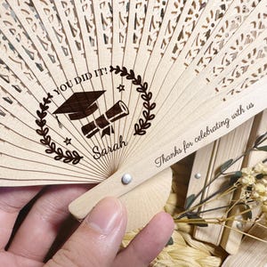 Könnte beinhalten: Ein Fächer aus Holz mit einem Design aus einem Abschluss-Hut und einem Diplom. Der Fächer ist mit dem Text "You Did It!" und "Thanks for celebrating with us" graviert.