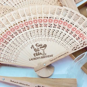 May include: Wooden folding fan with laser-cut floral and geometric patterns. The fan has a light brown color with red floral accents and the text "A Tini Bit Older LAUREN'S BIRTHDAY". The fan also has the text "Thank you for celebrating with me".