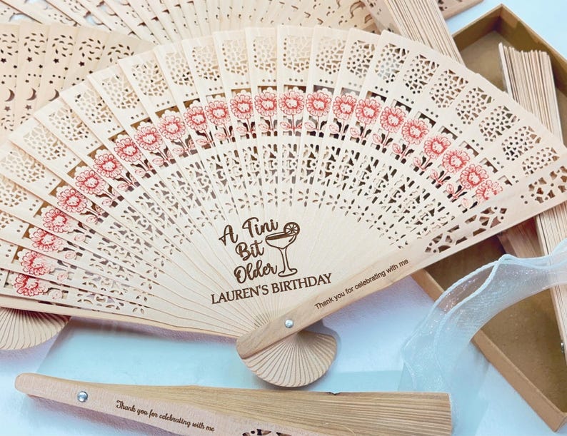 May include: Wooden folding fan with laser-cut floral and geometric patterns. The fan has a light brown color with red floral accents and the text "A Tini Bit Older LAUREN'S BIRTHDAY". The fan also has the text "Thank you for celebrating with me".