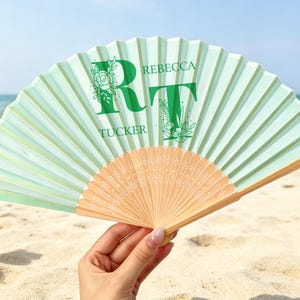 Puede incluir: Un abanico plegable de color verde claro con un marco de madera con recortes intrincados. El abanico muestra las letras R y T en verde oscuro, decoradas con arte lineal floral, junto con los nombres Rebecca y Tucker en texto verde oscuro.