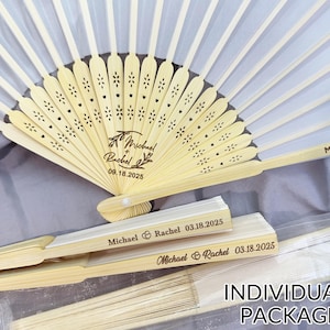 May include: White and beige folding fans with personalized text, including names and a date. The fans are made of wood and fabric, and are individually packaged. The text on the fan reads "Michael & Rachel 09.18.2025".