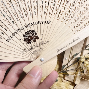 May include: A light-coloured wooden folding fan with detailed cut-out patterns. The fan is inscribed with the words "IN LOVING MEMORY OF" and "Sarah Wilson 1955-2025", along with a floral graphic. The phrase "Forever in our hearts" is also visible.