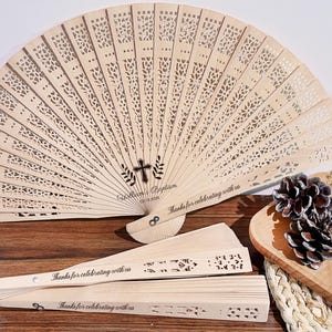 Puede incluir: Abanicos de madera de color marrón claro con intrincados diseños calados. Los abanicos muestran el texto "Thanks for celebrating with us" y "William's Baptism". También se ve una cruz en el abanico.