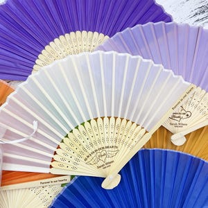 Puede incluir: Una colección de abanicos plegables coloridos en tonos morado, azul, naranja y amarillo. Algunos abanicos tienen el texto "Always in our hearts" y "Forever in our hearts" con un nombre y una fecha. Los abanicos son de papel y madera.