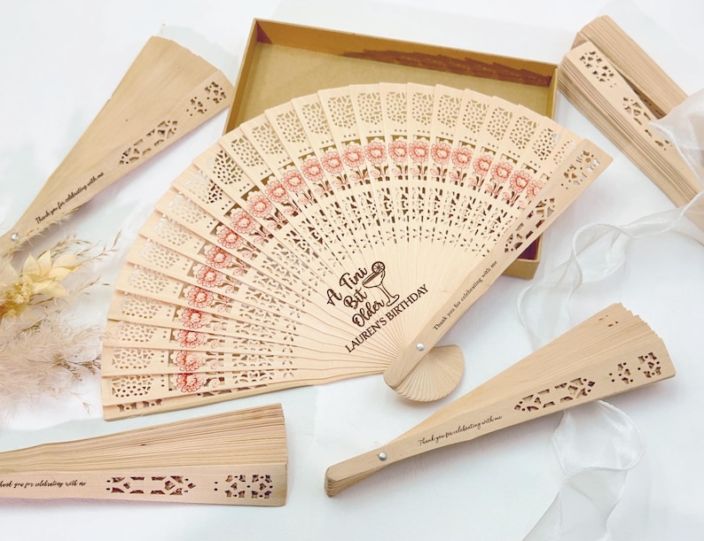 May include: Wooden hand fans with laser-cut designs and floral accents. The main fan has the text "A Tiny Bit Older, Lauren's Birthday". Other fans have the text "Thank you for celebrating with me".