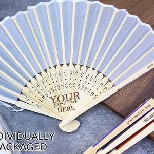 Puede incluir: Un abanico plegable blanco y azul claro con mango de madera. El abanico tiene el texto "YOUR Logo Text HERE" y "your custom text" impreso. El abanico está empaquetado individualmente.