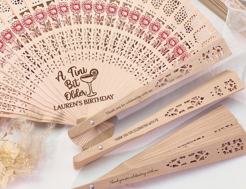 May include: Wooden fans with laser-cut designs and floral accents. One fan is open, displaying the text "A Tini Bit Older LAUREN'S BIRTHDAY." The other fans are closed, with the text "Thank you for celebrating with me."