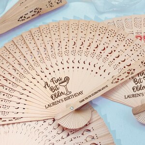 May include: Wooden hand fans with intricate cut-out designs featuring stars and crescent moons. The fans have the text "A Tini Bit Older LAUREN'S BIRTHDAY" and a cocktail illustration. Some fans have floral designs.