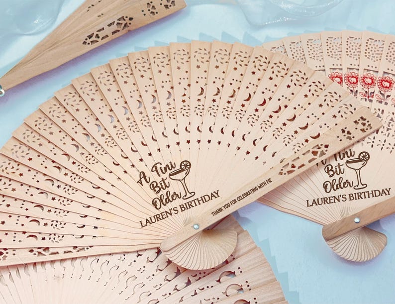 May include: Wooden hand fans with intricate cut-out designs featuring stars and crescent moons. The fans have the text "A Tini Bit Older LAUREN'S BIRTHDAY" and a cocktail illustration. Some fans have floral designs.