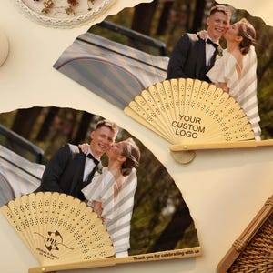 May include: Wooden hand fans with a printed photograph of a couple kissing. The fans have a decorative, openwork design and the text "YOUR CUSTOM LOGO" is printed on one fan. The other fan has the text "Michael & Rachel 03.18.2023".