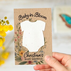Puede incluir: Tarjeta de invitación a un baby shower con el texto "Baby in Bloom" y "Christina's BABY SHOWER". La tarjeta presenta una forma de body blanco e ilustraciones florales en rosa, amarillo y azul sobre un fondo marrón.