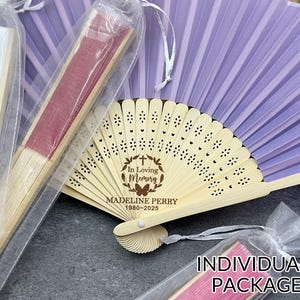 Puede incluir: Una colección de abanicos decorativos, algunos en envases transparentes. Un abanico está abierto, con un diseño plisado de color lila claro. El abanico abierto tiene un mango de madera con patrones florales cortados con láser y el texto "In Loving Memory Madeline Perry 1980-2025".