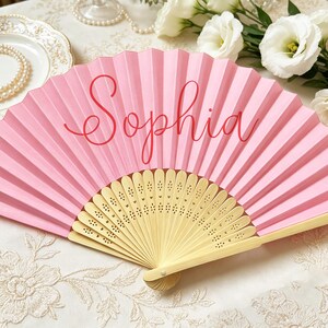 Puede incluir: Un abanico plegable de color rosa con el nombre Sophia escrito en letra cursiva roja en el centro. El abanico cuenta con una estructura de madera clara con intrincados patrones florales cortados a láser en la base, un accesorio delicado.