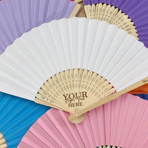 Puede incluir: Abanico de papel blanco con detalles de madera, con el texto "YOUR LOGO/TEXT HERE". Está rodeado de otros abanicos en varios colores, incluyendo morado, rosa y azul. El texto "Your custom text" también es visible.