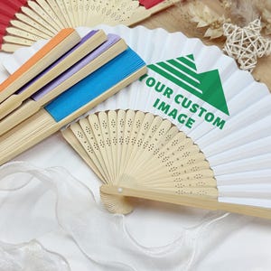 Puede incluir: Una colección de abanicos plegables con mangos de bambú. Los abanicos presentan varios colores, incluyendo rojo, naranja, morado y azul. Un abanico tiene el texto "YOUR CUSTOM IMAGE" impreso. Los abanicos están dispuestos sobre una superficie blanca.