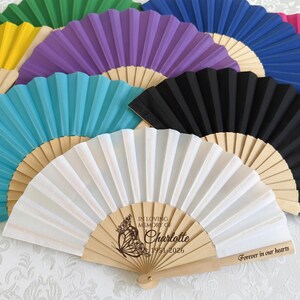 Puede incluir: Una colección de abanicos de papel coloridos con mangos de madera. Los abanicos son de varios colores, incluyendo blanco, negro, azul, morado y verde. Un abanico tiene el texto "In Loving Memory of Charlotte 1951-2026" y "Forever in our hearts."