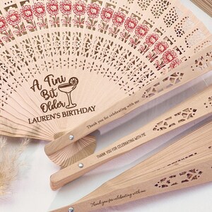 May include: Wooden fans with laser-cut designs and floral accents. One fan is open, displaying the text "A Tini Bit Older LAUREN'S BIRTHDAY." The other fans are closed, with the text "Thank you for celebrating with me."