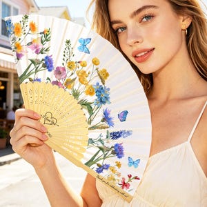 Puede incluir: Un abanico plegable con base color crema y un estampado floral vibrante con flores silvestres amarillas, azules y moradas y pequeñas mariposas azules. El mango de madera incluye el texto I Love You y Thank you for celebrating with us.