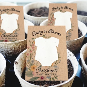 Può includere: Sono esposti diversi favori per baby shower, ciascuno con un biglietto di carta kraft con un ritaglio di tutina bianca. Le carte sono decorate con illustrazioni floreali e il testo "Baby in Bloom" e "Christina's BABY SHOWER."