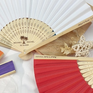 Puede incluir: Varios abanicos decorativos de diferentes colores. Un abanico es blanco, otro es beige con un diseño de palmera y los nombres "Michael & Rachel" y la fecha "03.18.2025". Otros abanicos son naranjas, morados y rojos. Los abanicos son de papel y bambú.