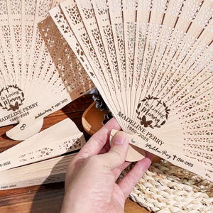 Puede incluir: Abanicos de madera clara con intrincados diseños calados. Los abanicos presentan el texto "In Loving Memory" y el nombre "MADELINE PERRY 1980-2025". Tienen un diseño delicado y aireado.