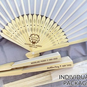 May include: White and beige folding fans with a floral design and the text "In Loving Memory Madeline Perry 1980-2025". The fans are individually packaged and made of paper and wood.
