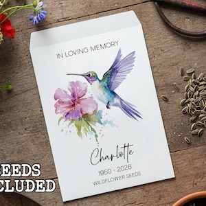Peut inclure: Sachet de graines blanc avec un motif aquarelle d'un colibri et d'une fleur. Le texte indique "In Loving Memory" et "Charlotte 1950-2026 Wildflower Seeds". Les mots "Seeds Included" sont également visibles.