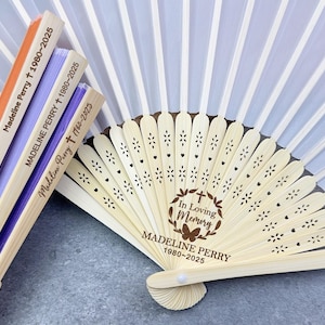 Puede incluir: Abanicos conmemorativos personalizados con el nombre "Madeline Perry" y las fechas 1980-2025. Los abanicos de madera y papel también muestran el texto "In Loving Memory". Los abanicos son de color naranja, morado y blanco.