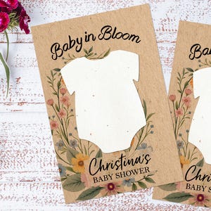 Puede incluir: Dos invitaciones para baby shower con diseño floral. Las invitaciones son de cartulina marrón con un recorte en forma de body. El texto "Baby in Bloom" y "Christina's Baby Shower" está impreso en las tarjetas.