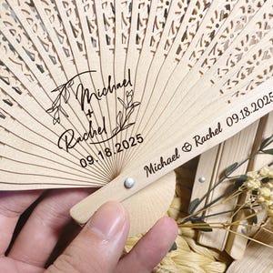 Puede incluir: Un abanico de madera clara con intrincados diseños calados. El abanico está personalizado con los nombres "Michael + Rachel" y la fecha "09.18.2025". Los nombres y la fecha están impresos en negro. El abanico está parcialmente abierto.
