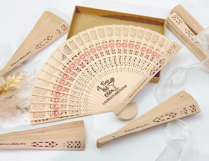 May include: Wooden fans with intricate cut-out designs and floral patterns in red. The central fan features the text "A Tiny Bit Older, Lauren's Birthday". Several fans are displayed, some open and some closed, with a gift box.