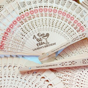 May include: A collection of light-colored wooden fans with intricate cut-out designs. Some fans feature red floral patterns and the text "A Tini Bit Older LAUREN'S BIRTHDAY". Other fans have the text "Thank you for celebrating with me".