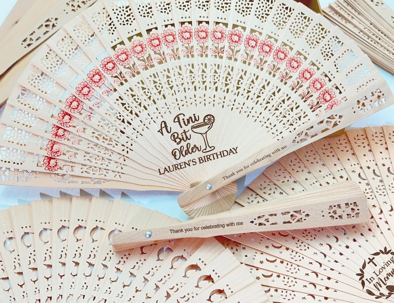 May include: A collection of light-colored wooden fans with intricate cut-out designs. Some fans feature red floral patterns and the text "A Tini Bit Older LAUREN'S BIRTHDAY". Other fans have the text "Thank you for celebrating with me".