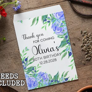 May include: A white seed packet with blue floral watercolor design and the text "Thank you for coming Olivia's 60th Birthday 12.28.2028". The words "Seeds Included" are at the bottom. The packet is on a wooden surface.