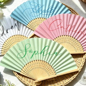 Puede incluir: Cuatro abanicos plegables con varillas de madera clara y papel plisado en azul pastel, blanco, rosa y verde claro. Cada abanico presenta un nombre escrito en elegante letra cursiva: Elizabeth en azul, Audrey en negro, Catherine en rosa y Sophia en verde.