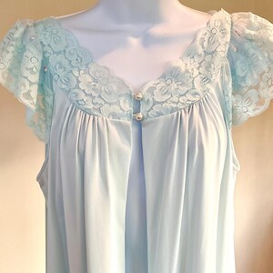May include: A light blue vintage nightgown with a delicate lace trim around the neckline and sleeves. The nightgown features a V-neckline and is fastened with two small pearl-like buttons. The fabric has a soft, flowing appearance.