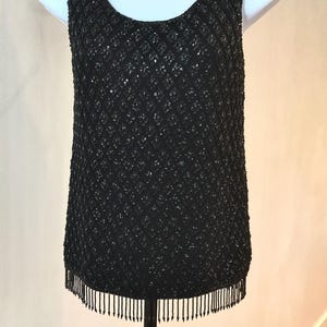 May include: A sleeveless black tank top featuring an all-over diamond-shaped pattern of small, reflective black sequins. The garment has a wide scoop neckline and a straight hemline finished with a row of delicate, dangling black beaded fringe.