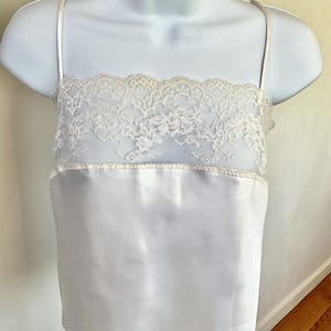 Pu&ograve; includere: Una camisole bianca con delicate spalline sottili. La parte superiore presenta un'ampia fascia di pizzo color crema. Il tessuto liscio, simile al raso, della camisole cade delicatamente. L'indumento &egrave; esposto su un manichino bianco.