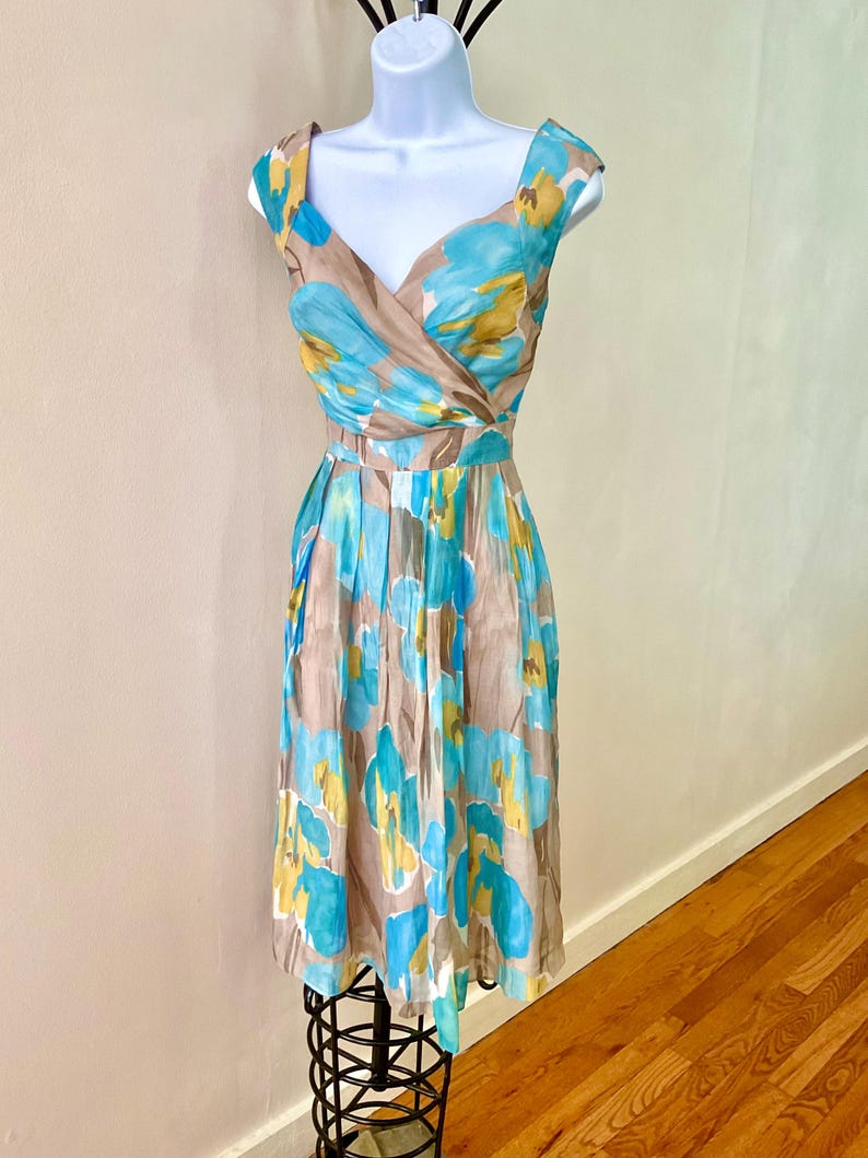 Puede incluir: Vestido sin mangas con escote en V y falda a la rodilla. El vestido presenta un estampado floral en tonos azules, amarillos y marrones. El vestido se exhibe en un maniqu&iacute; blanco. El vestido tiene cintura entallada y falda plisada.