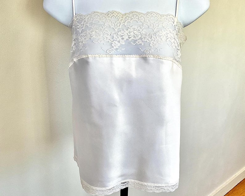 Pu&ograve; includere: Una camisole di raso bianco con delicate finiture in pizzo sullo scollo e sull'orlo. La camisole ha sottili spalline e un design semplice ed elegante. I dettagli in pizzo aggiungono un tocco di fascino vintage.