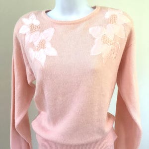 May include: A pale pink sweater with long sleeves and a crew neck. The sweater has white embroidered flowers on the shoulders.