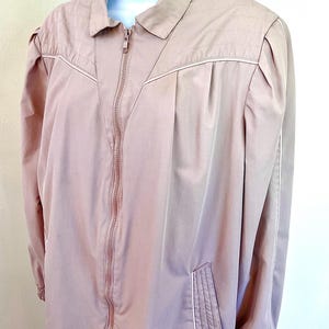 May include: A dusty rose zip-up jacket featuring a pointed collar, long sleeves with elastic cuffs, and white piping accents along the shoulders and pockets. The garment includes a full-length front zipper and quilted detailing on the upper chest.