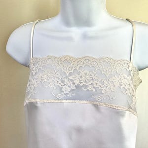 Pu&ograve; includere: Camicia di raso azzurro con sottili spalline. La parte superiore presenta un pannello in pizzo color crema sul petto. Il pizzo ha un motivo floreale. La camicia &egrave; esposta su un manichino bianco.