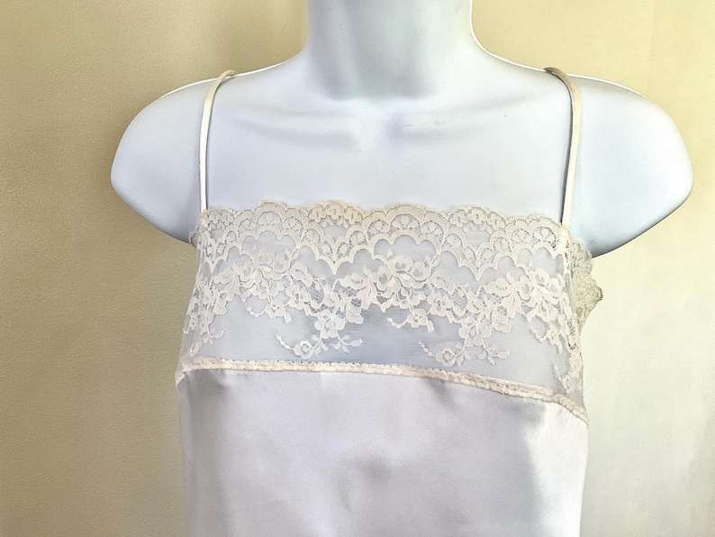 Pu&ograve; includere: Camicia di raso azzurro con sottili spalline. La parte superiore presenta un pannello in pizzo color crema sul petto. Il pizzo ha un motivo floreale. La camicia &egrave; esposta su un manichino bianco.