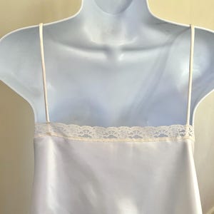 Pu&ograve; includere: Abito sottoveste in raso bianco con sottili spalline e un bordo in pizzo color crema lungo lo scollo. L'abito &egrave; esposto su un manichino bianco, che ne mette in risalto l'eleganza semplice e la consistenza liscia. Lingerie classica.