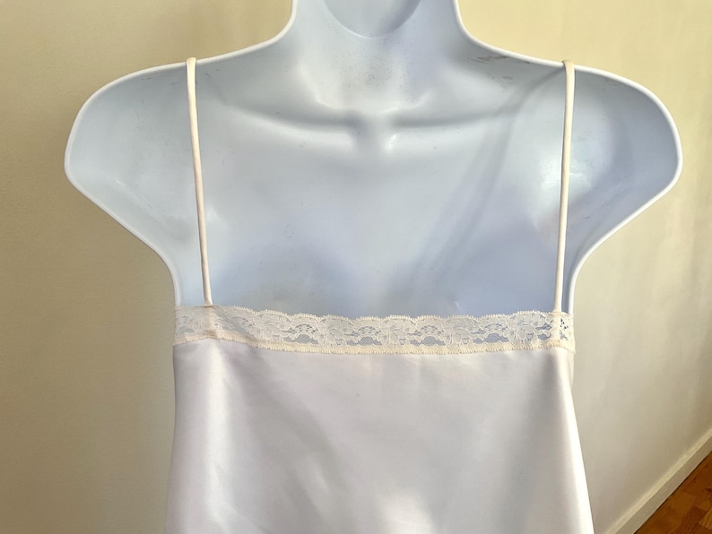Pu&ograve; includere: Abito sottoveste in raso bianco con sottili spalline e un bordo in pizzo color crema lungo lo scollo. L'abito &egrave; esposto su un manichino bianco, che ne mette in risalto l'eleganza semplice e la consistenza liscia. Lingerie classica.