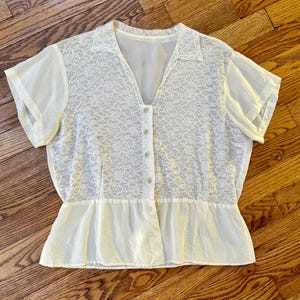 Puede incluir: Una blusa blanca de encaje con botones en la parte delantera y mangas cortas. La blusa tiene un dobladillo con volantes y una superposición transparente.