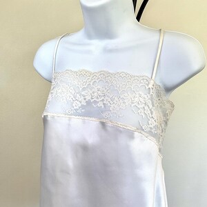 Pu&ograve; includere: Un abito sottoveste in raso bianco con delicate spalline sottili. Il corpetto presenta un'ampia fascia di pizzo color crema. L'abito &egrave; esposto su un manichino bianco, mostrando il suo design elegante e la silhouette fluida.