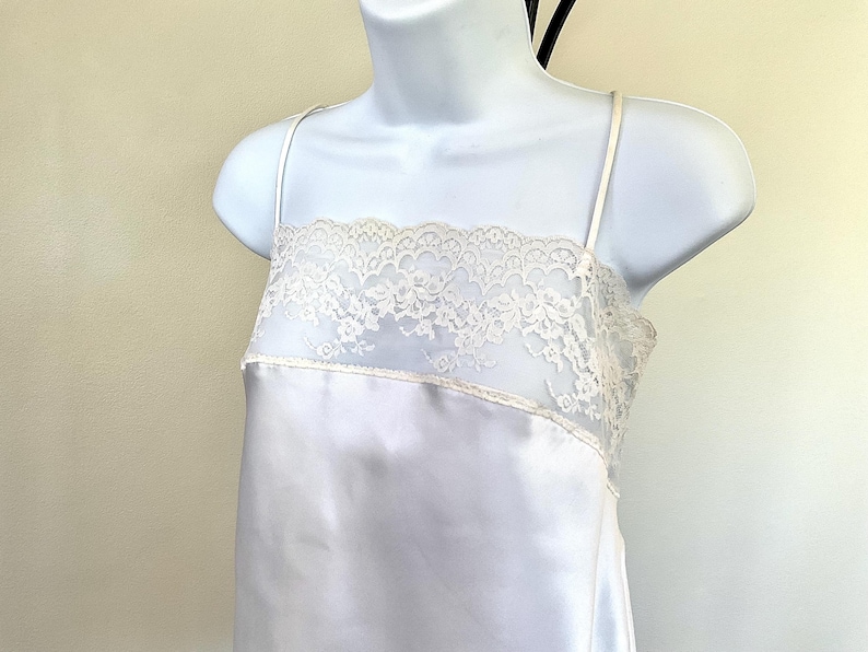 Pu&ograve; includere: Un abito sottoveste in raso bianco con delicate spalline sottili. Il corpetto presenta un'ampia fascia di pizzo color crema. L'abito &egrave; esposto su un manichino bianco, mostrando il suo design elegante e la silhouette fluida.